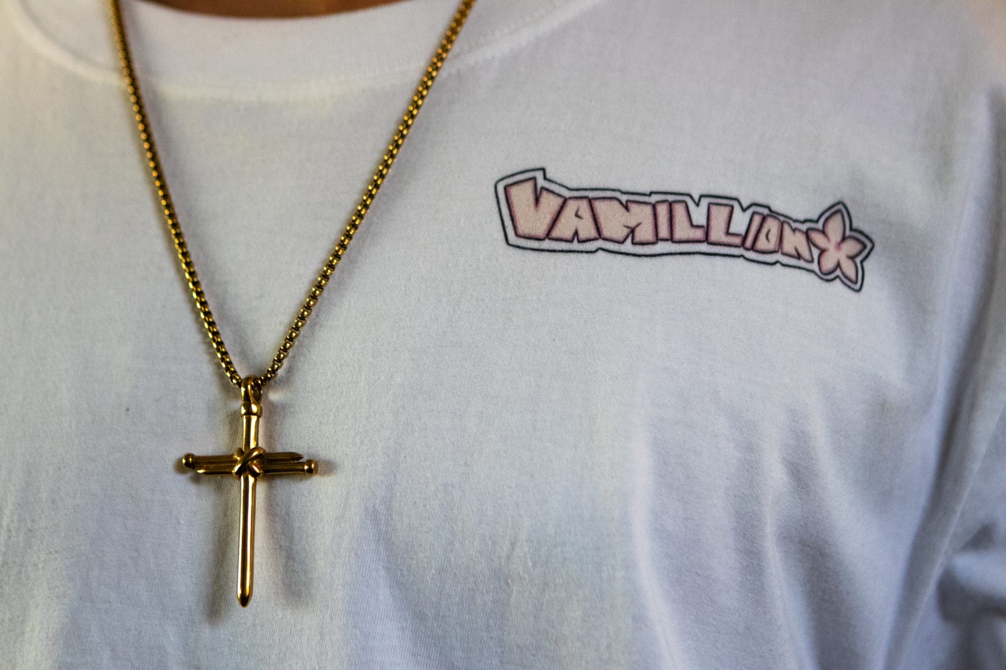 A gold nail cross necklace over a white tee with a small VAMILLION chest logo
